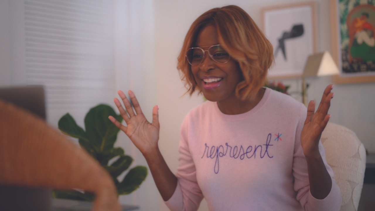 A smiling woman wears glasses and a pink sweater with the word Represent on the front