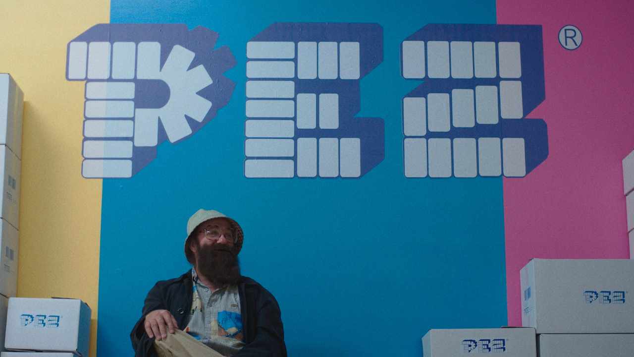 a man with a beard and bucket hat sitting in front of a Pez logo mural