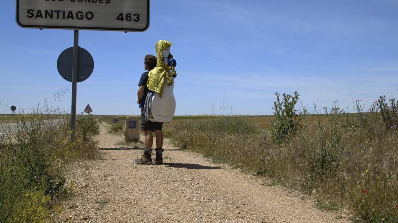 a man with a cello on his back standing on a dirt road next to a sign post