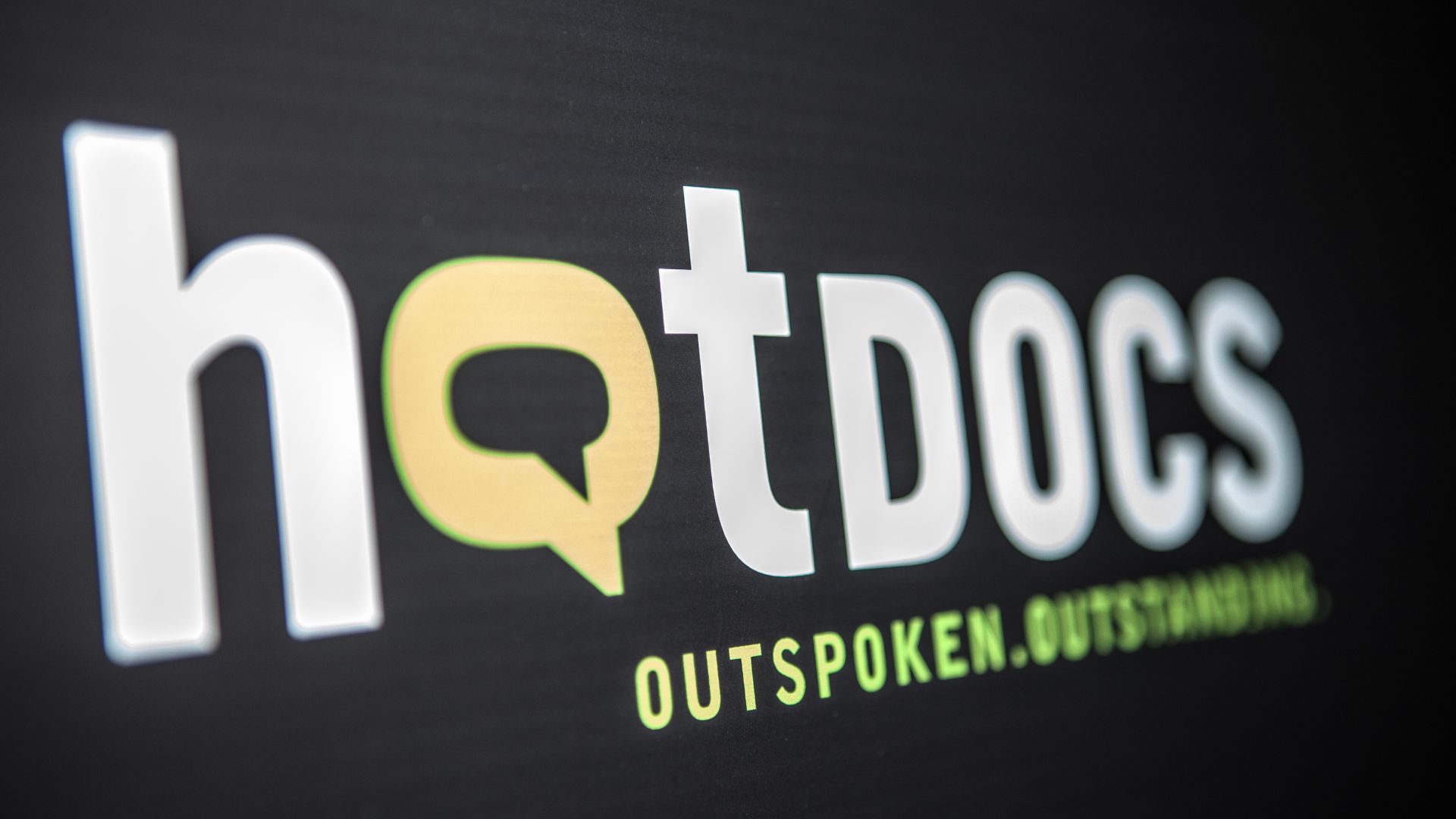 White and green Hot Docs logo with tagline that reads 'Outspoken. Outstanding.' against a black background.