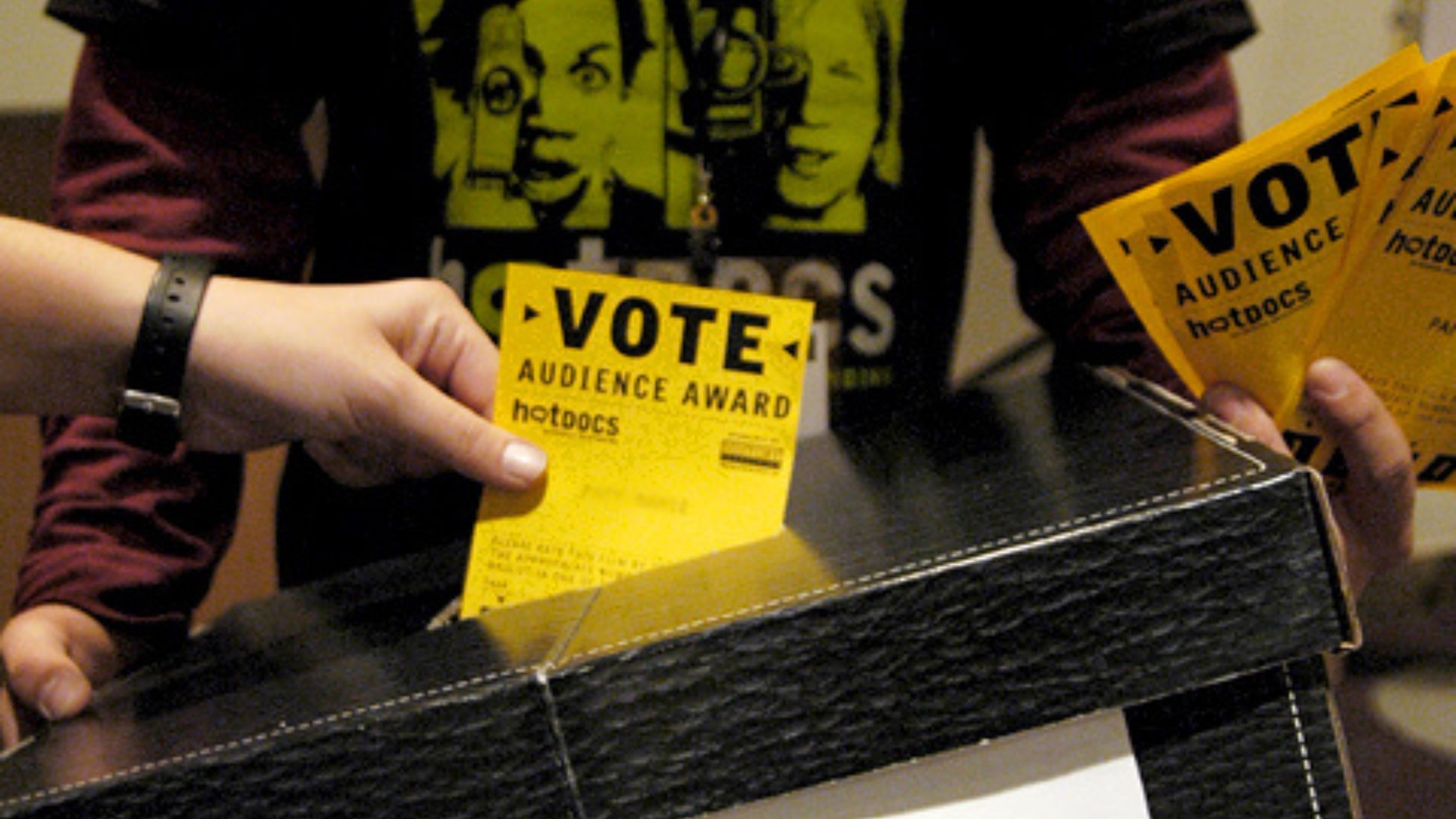 A person inserting a yellow Audience Vote card into a black ballot box.